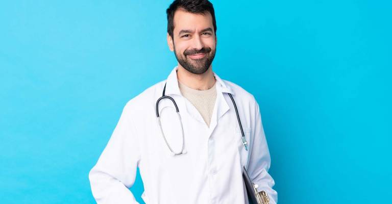 Jonge blanke man die een doktersjas draagt, stethoscoop om heeft ​​en een map vasthoudt op een blauwe achtergrond